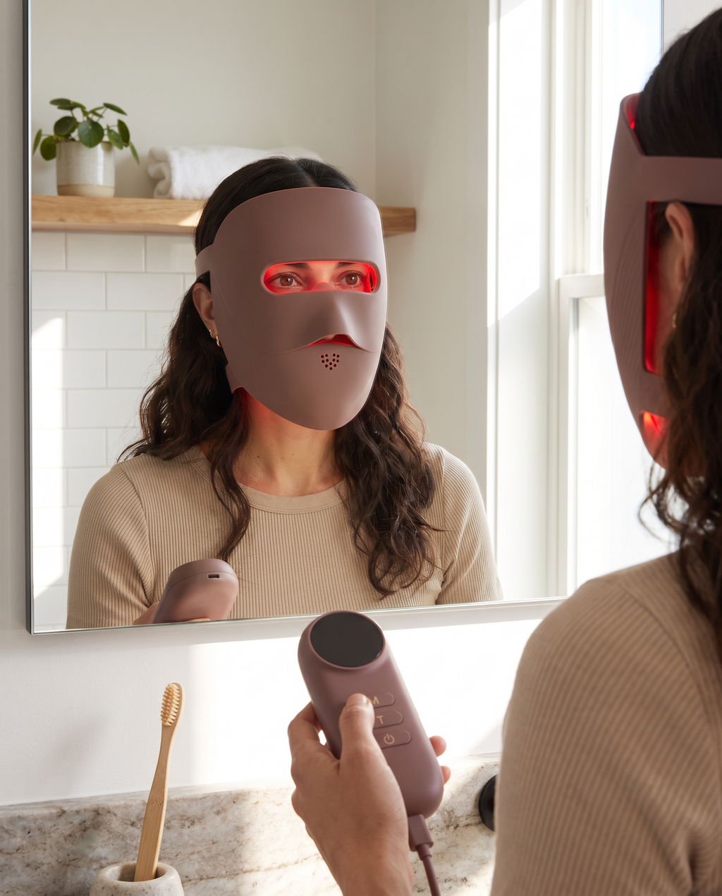 Clinaray LED mask in Brown worn at a bathroom mirror, red mode on, eyes open through the eye window, controller in hand. Close your eyes or use shields if the light bothers you.